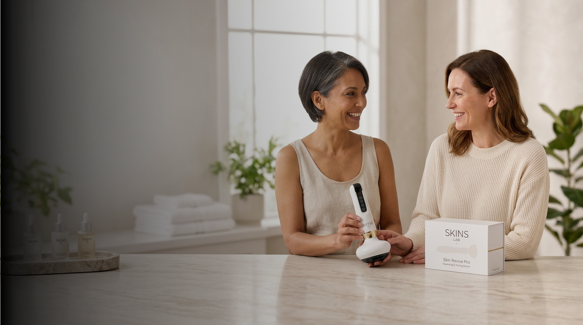 Two women sitting at a marble counter in a bright bathroom setting, smiling and talking. One holds a white handheld beauty device, and the other points to a white SHINS box on the counter.